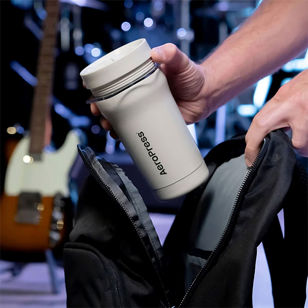 Person holding a beige Aeropress coffee mug with a blurred background of musical instruments.