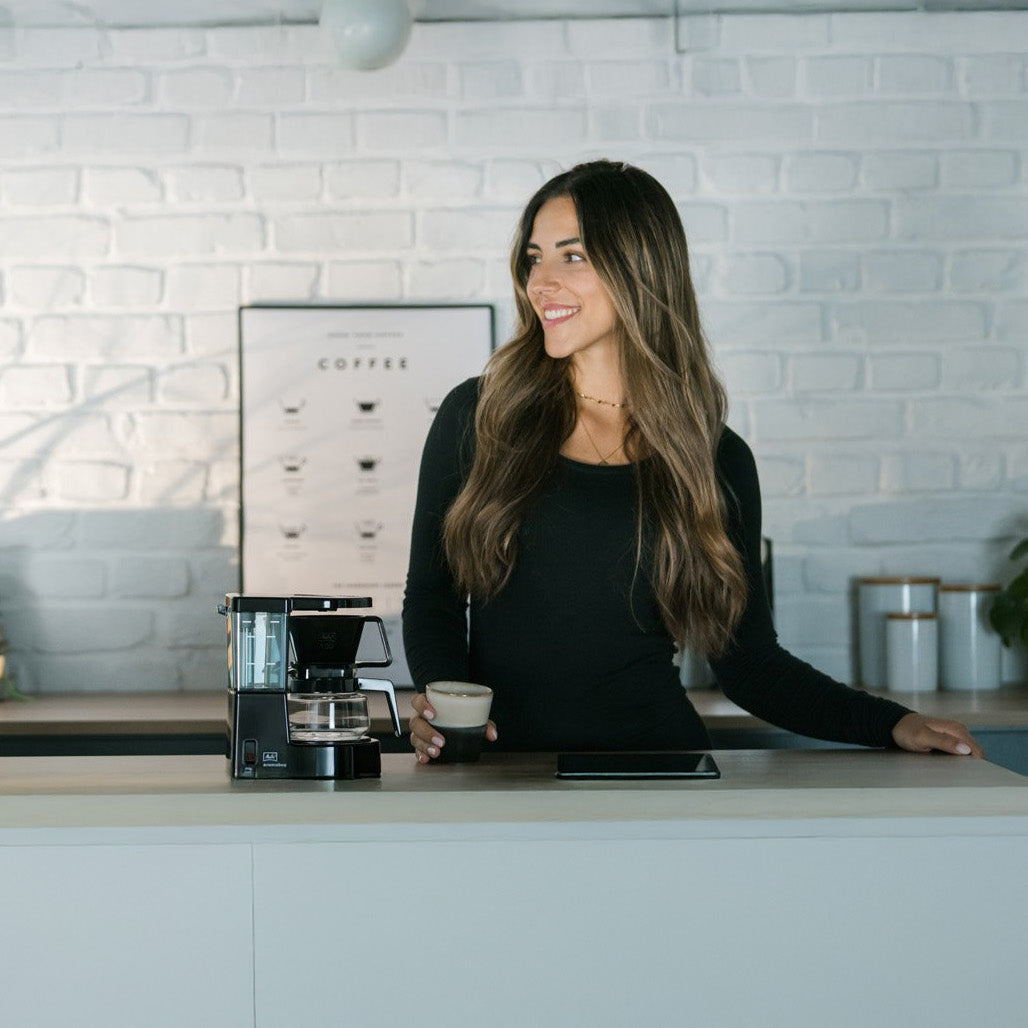 Woman standing in a kitchen with a coffee maker and a cup.
