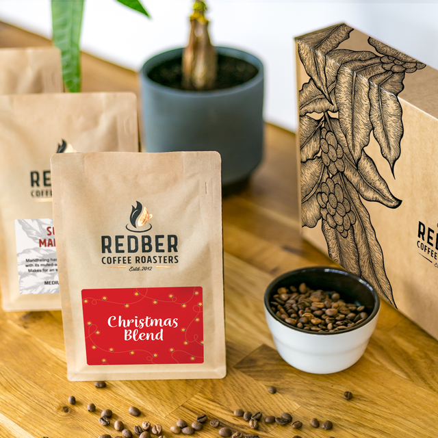 Redber Coffee Roasters Christmas Blend package on a wooden surface with coffee beans and a plant in the background.
