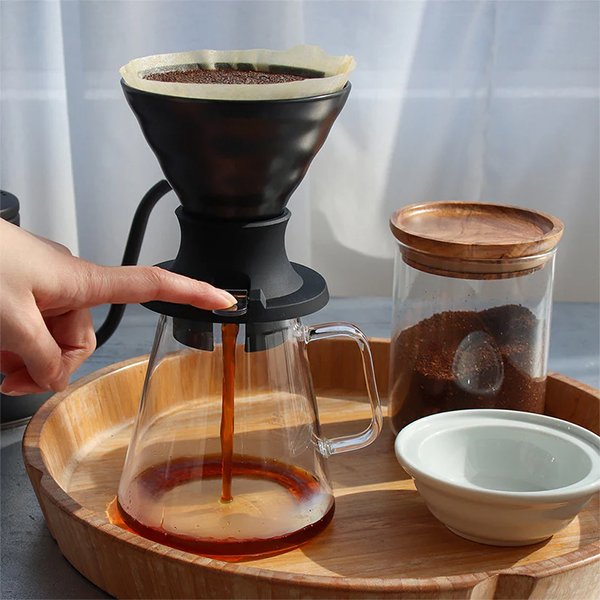 Hand pouring coffee from a pour-over coffee maker onto a wooden tray with a glass carafe, wooden container, and small white bowl.