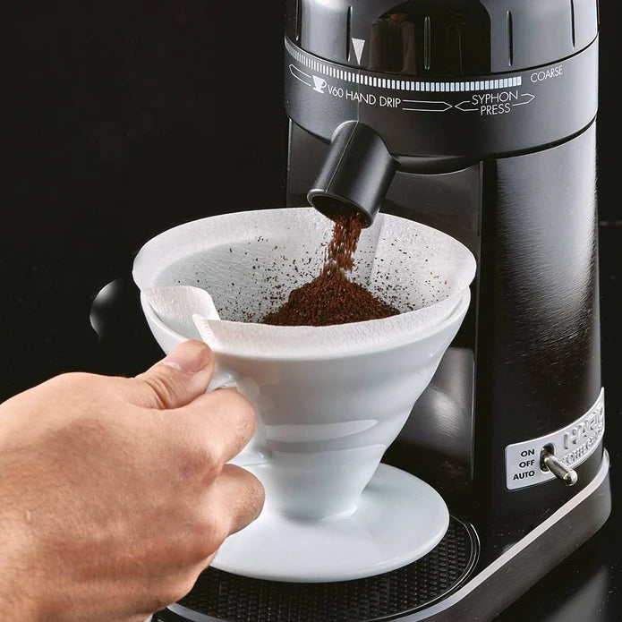 Hand pouring coffee grounds into a white filter over a black coffee machine.