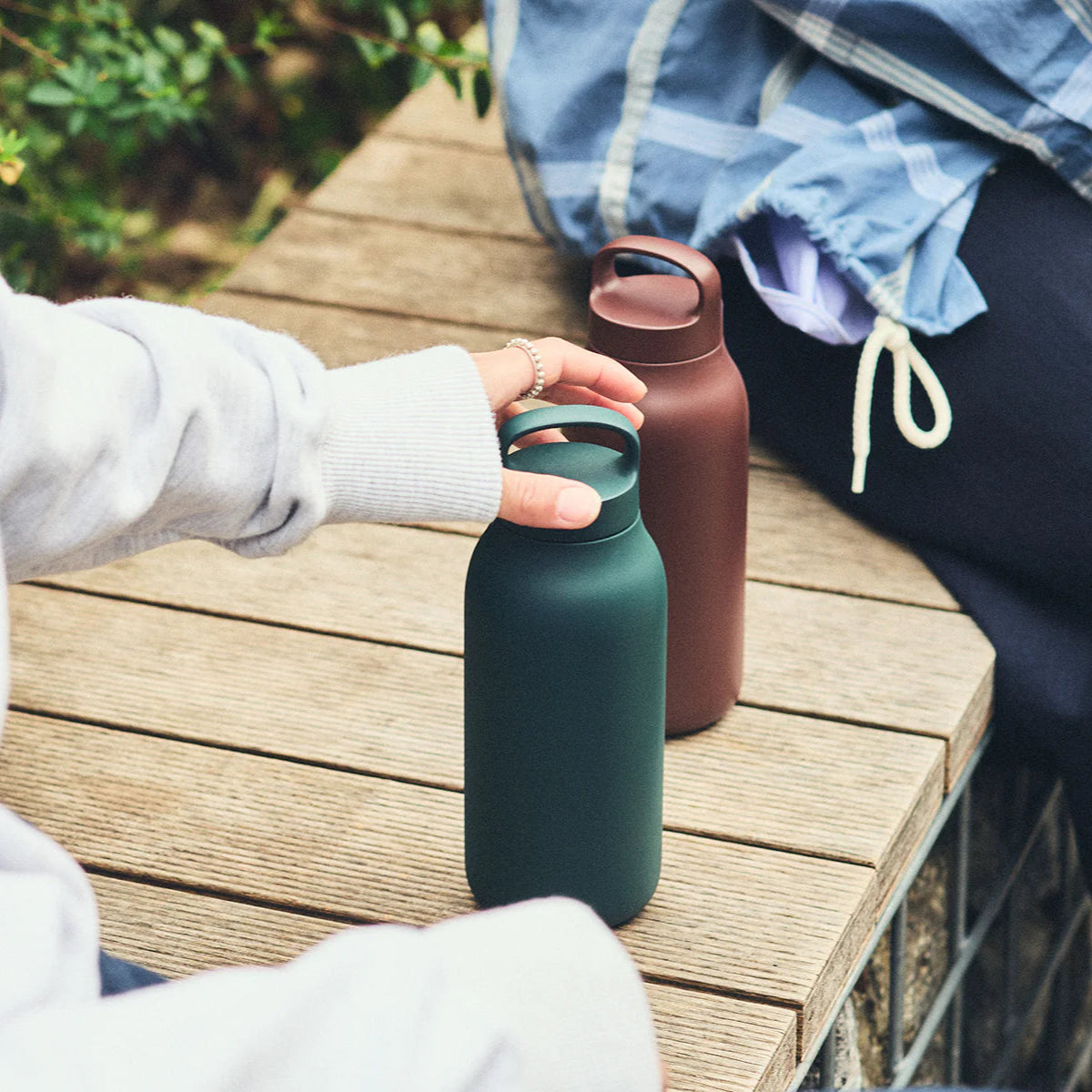 Two people with green and brown water bottles on a wooden table outdoors.