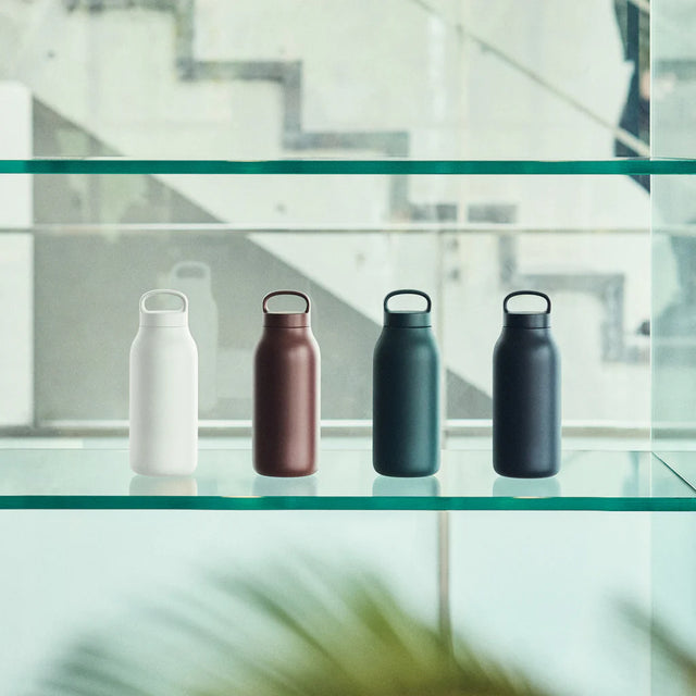 Four metal water bottles on a glass shelf with a blurred indoor background