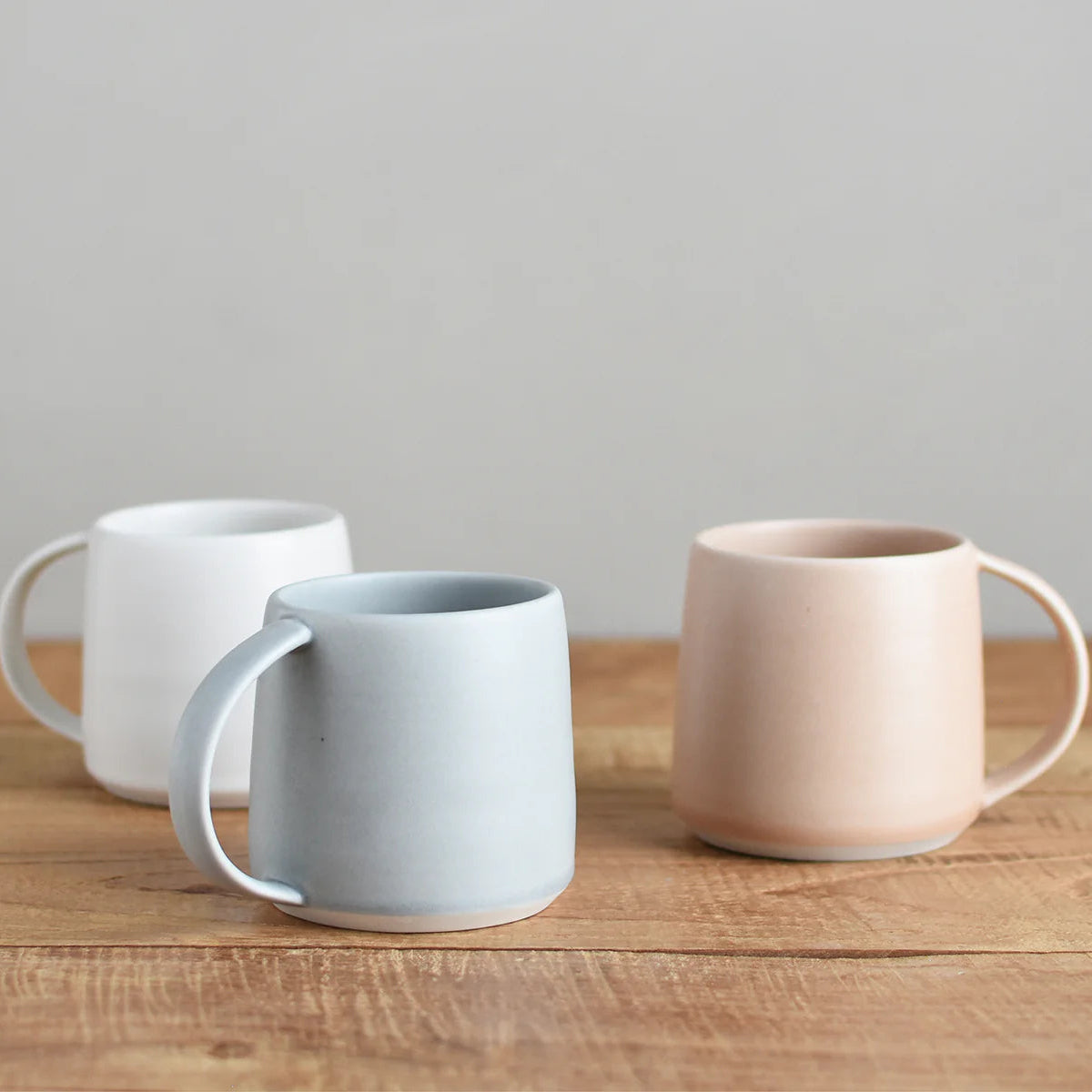 Three ceramic mugs in white, blue, and pink on a wooden surface with a gray background