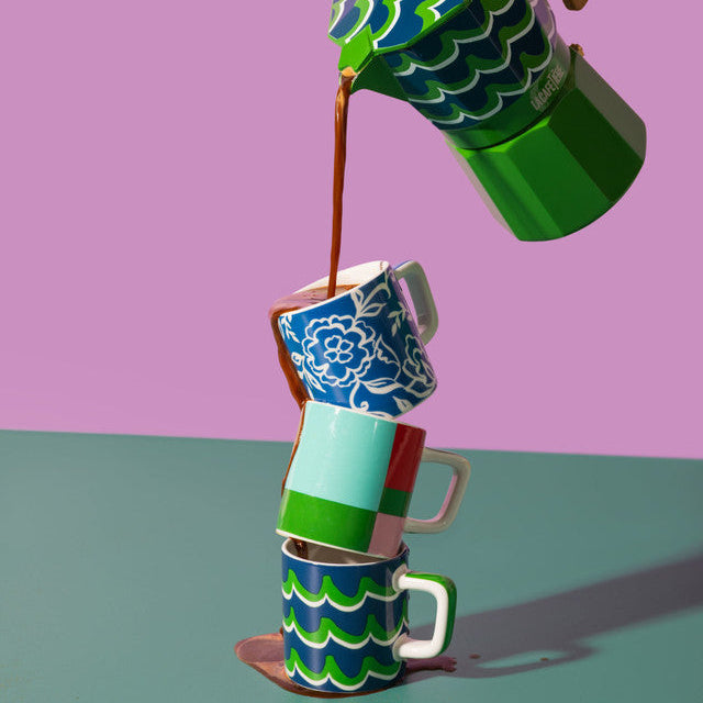 Colorful coffee press pouring coffee into a stack of patterned cups against a purple and green background.