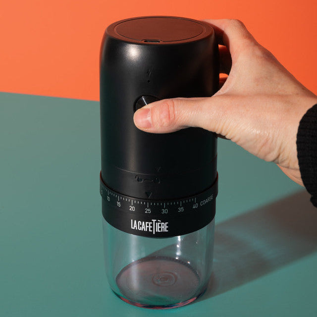 Hand holding a black and clear coffee grinder with 'La Cafetiere' branding against an orange and green background.