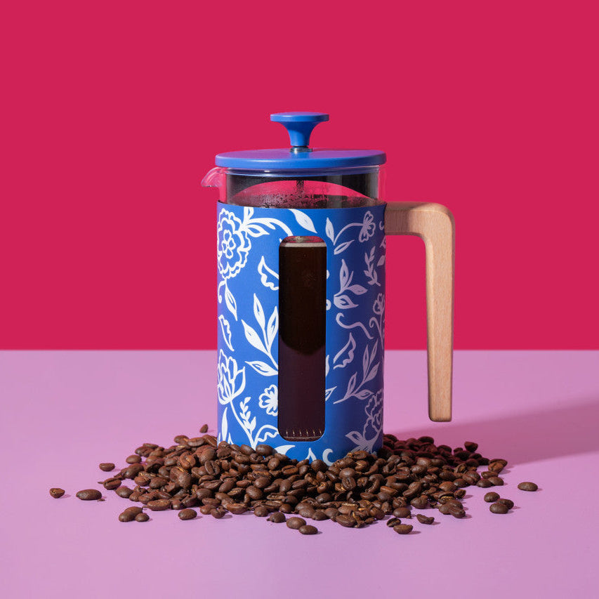 Blue and white patterned French press with a wooden handle on a pink background with coffee beans around it.