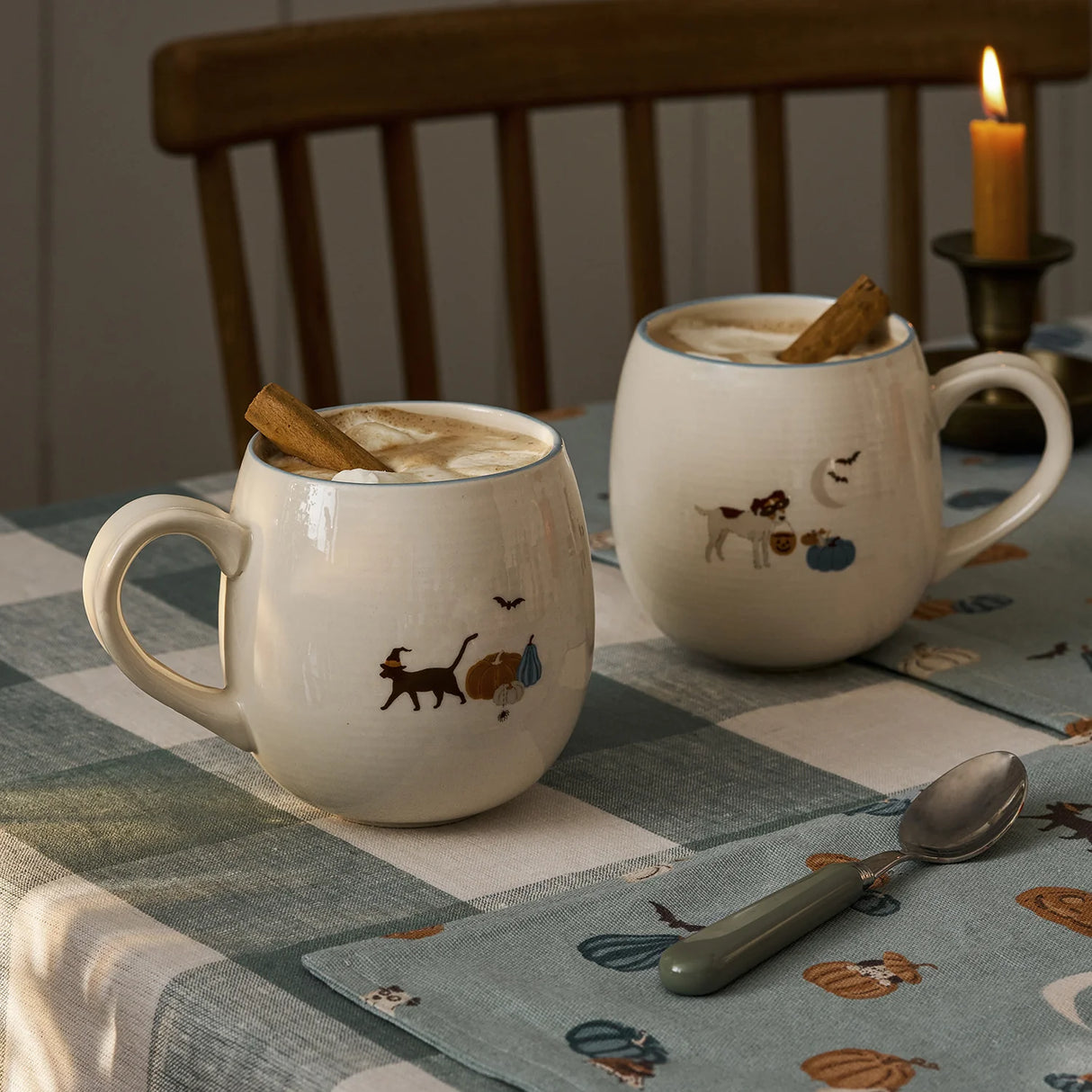 Two ceramic mugs with animal illustrations on a checkered tablecloth with a candle in the background.