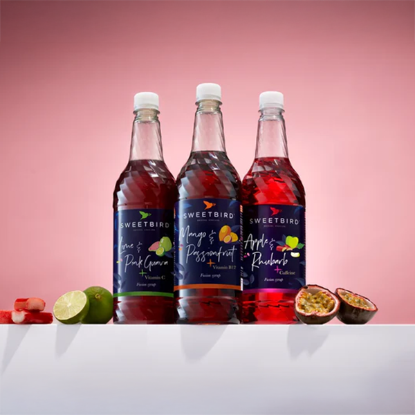 Three bottles of Sweetbird syrup with fruit slices on a pink background
