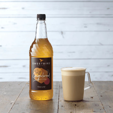 Bottle of Sweetbird gingerbread syrup next to a cup of coffee on a wooden surface.