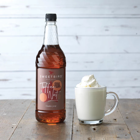 Bottle of Sweetbird Toffee Nut syrup next to a cup of hot chocolate with whipped cream on a wooden table.