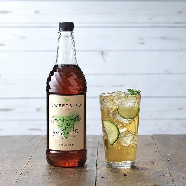 Bottle of Sweetbird syrup next to a glass of iced tea with lime and cucumber slices on a wooden surface.
