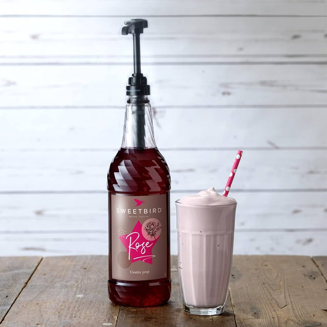 Bottle of Sweetbird Rose syrup next to a glass of pink milkshake on a wooden surface.