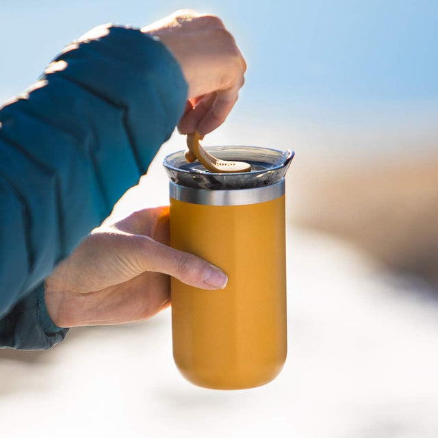 Person holding a yellow insulated tumbler with a blue sleeve against a blurred outdoor background