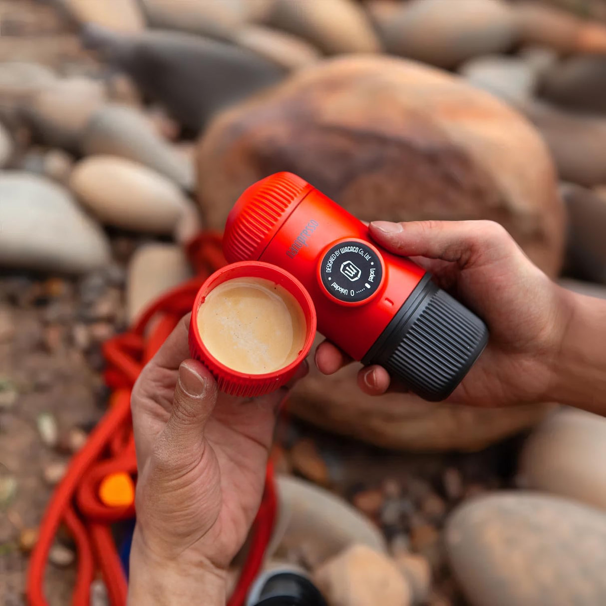 Red camping coffee maker with a lid open, showing coffee grounds, held by a person against a rocky background.