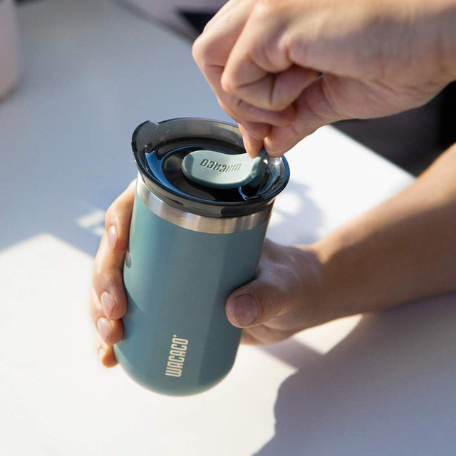 Person holding a teal travel mug with a black lid, opening it on a light surface. Wacaco Octaroma Lungo Vacuum Insulated Mug 300ml - Cadet Blue