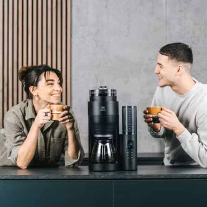 Two people in a kitchen with coffee makers and mugs.