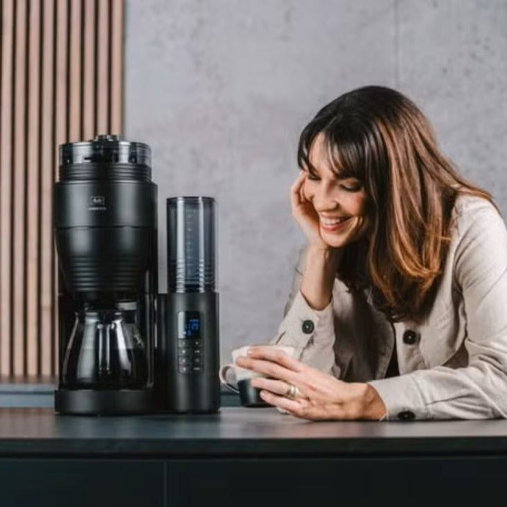 Woman sitting at a table with coffee-making equipment in a kitchen setting