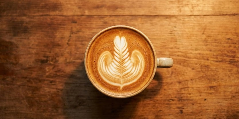 Top-down view of a freshly made flat white coffee featuring intricate rosetta latte art, sitting on a warm, textured wooden table.