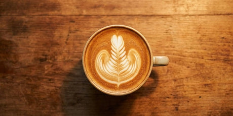 Top-down view of a freshly made flat white coffee featuring intricate rosetta latte art, sitting on a warm, textured wooden table.