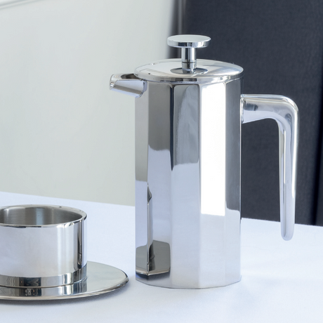 A polished stainless steel French press coffee maker with a matching metal cup and saucer on a white table, set against a neutral indoor background.