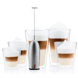A Bodum SCHIUMA Milk Frother Whisk in stainless steel, with a handheld design and a flexible stainless-steel wand, displayed alongside glasses of frothy beverage.