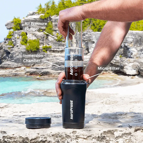Person holding an AeroPress coffee maker with labeled parts against a natural background.