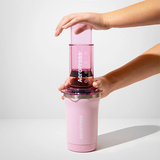 Pink AeroPress coffee maker being held by a person on a light gray background