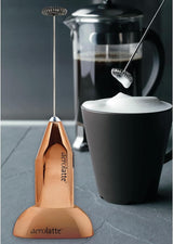 Aerolatte frother with a French press and cup in the background on a gray surface.