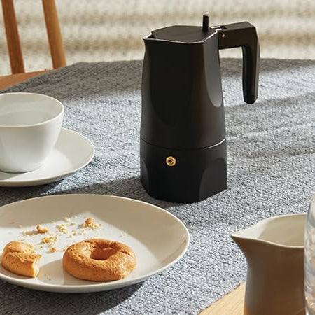 Black coffee maker on a table with a white cup, saucer, and plate with pastries.