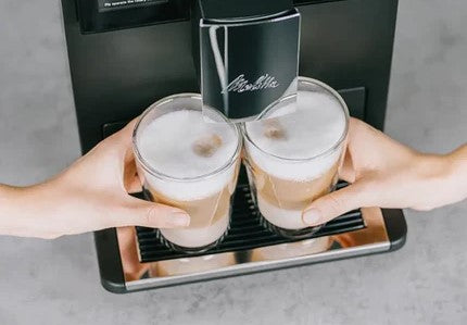 Two glasses of cappuccino being held up in front of a coffee machine.