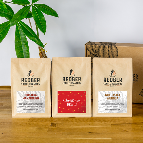 Three Redber Coffee Roasters coffee bags on a wooden surface with a plant in the background.