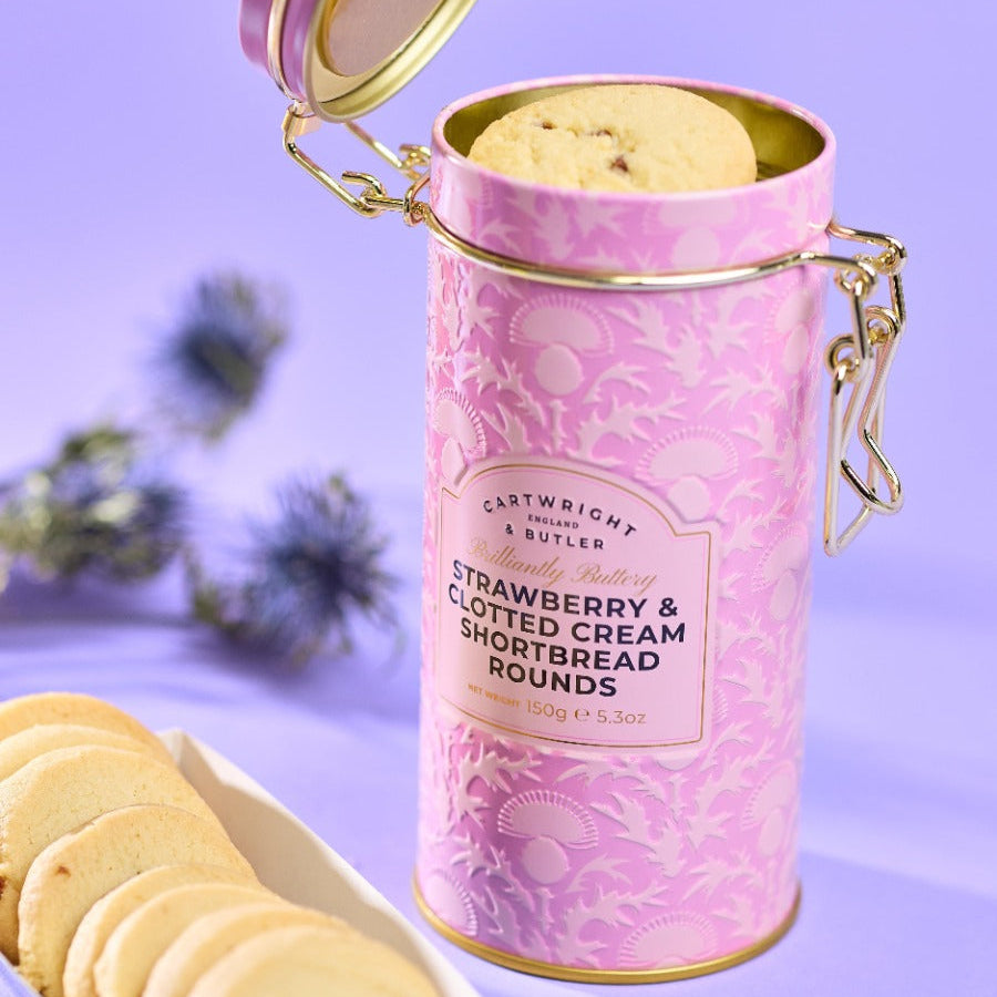 Pink cylindrical container of strawberry & clotted cream shortbread rounds with cookies on a plate and blurred plants in the background.