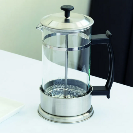 A glass French press with a stainless steel lid, plunger, and base, featuring a black handle and black top knob, sitting on a white table.