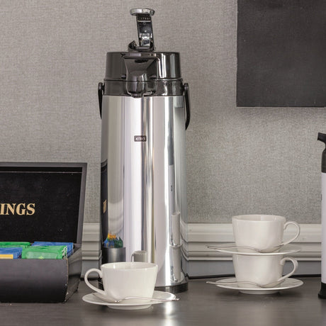 A countertop coffee station setup featuring a tall stainless steel coffee dispenser, a smaller white and black thermal carafe, stacked white cups and saucers, a black tea box with TWININGS branding, and a decorative lamp against a gray wall.
