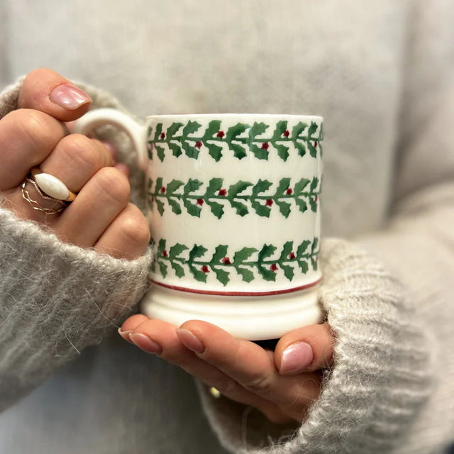 Person holding a mug with holly design, wearing a gray sweater.