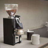Coffee grinder with coffee beans on a countertop with mugs.