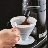 Hand pouring coffee grounds into a white filter over a black coffee machine.