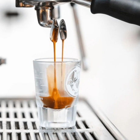Espresso being poured into a clear glass with a logo, likely from a professional espresso machine.