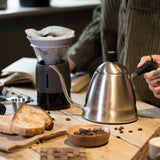 Person making coffee using a pour-over setup on a wooden table with bread and coffee beans.