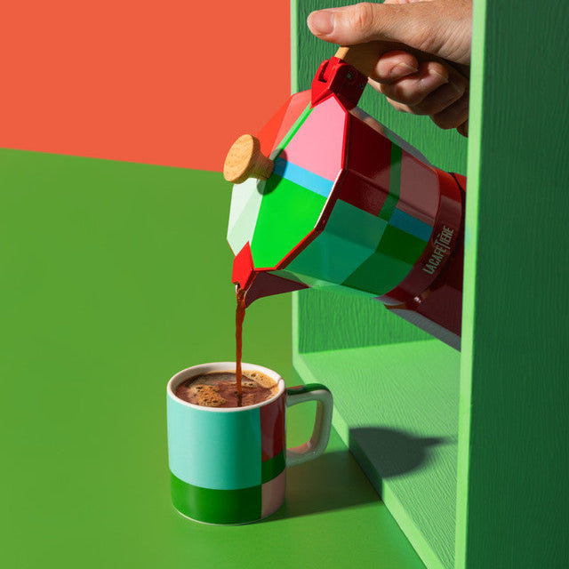 Colorful coffee pot pouring coffee into a mug on a green shelf with a red background