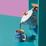 Person pouring coffee from a white and blue patterned coffee pot into a matching blue mug on a teal and pink background.