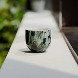 Marble-patterned ceramic cup on a white surface with blurred greenery in the background