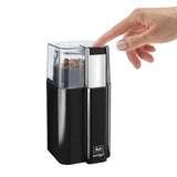 A hand pressing the button on the Melitta AMIGO Electric Blade Coffee Grinder, showing coffee beans inside the transparent lid and the grinder's sleek black and chrome design.