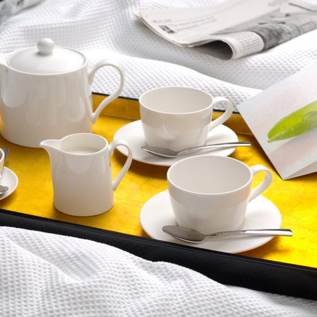 White ceramic tea and coffee set arranged on a yellow tray, including a teapot, cups with saucers, a small creamer pitcher, and spoons, styled on a bed with white textured bedding and a folded newspaper.