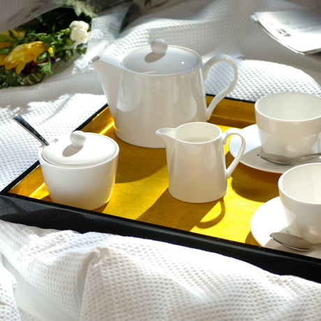 White ceramic tea and coffee serving set arranged on a black tray with a gold center, placed on white textured bedding with a newspaper and flowers nearby.