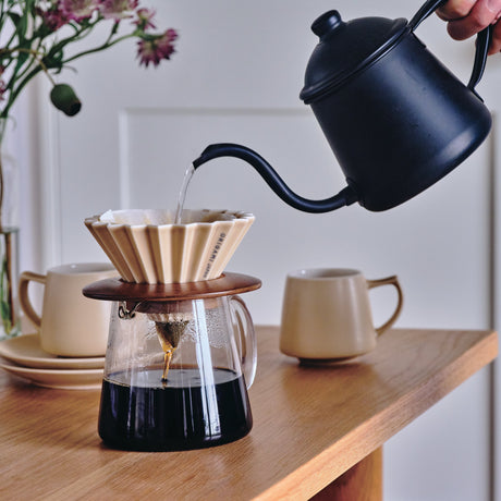 Person pouring coffee from a blue kettle into a filter on a wooden table with a vase of flowers in the background.