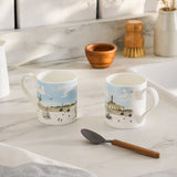 Two mugs with scenic designs on a marble countertop with a spoon and wooden items.