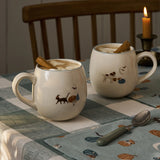 Two ceramic mugs with animal illustrations on a checkered tablecloth with a candle in the background.