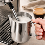 Person using a milk frothing pitcher with an espresso machine on a countertop.